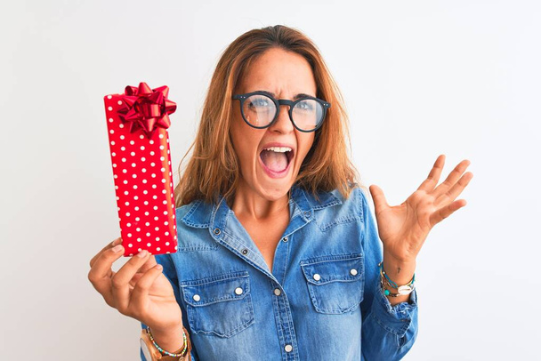 Young beautiful redhead woman wearing glasses and holding birthday present very happy and excited, winner expression celebrating victory screaming with big smile and raised hands - Valokuva, kuva