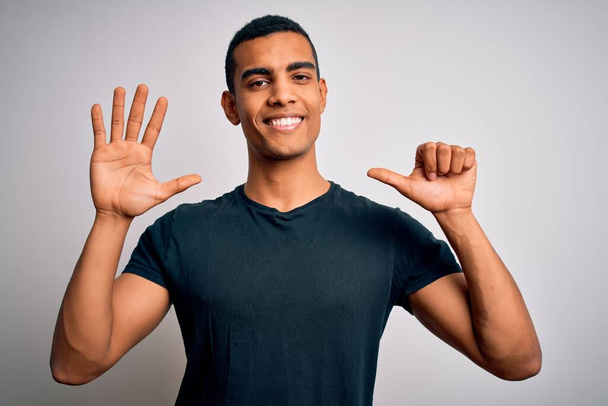 Nuori komea afrikkalainen amerikkalainen mies päällään rento t-paita seisoo valkoinen tausta näyttää ja osoittaa sormilla numero kuusi hymyillen itsevarma ja onnellinen
. - Valokuva, kuva