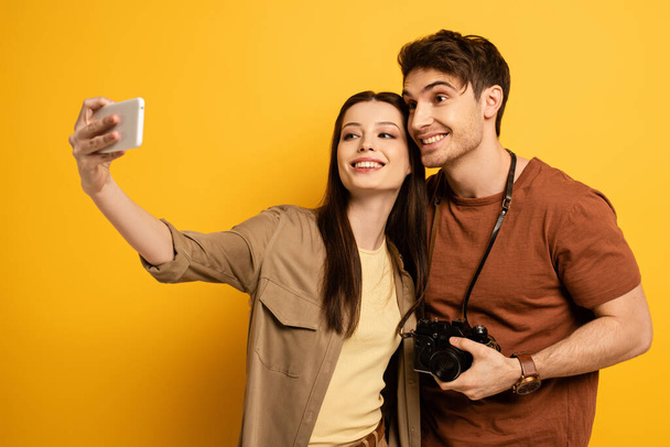 couple of happy tourists with photo camera taking selfie on smartphone on yellow - Foto, Imagen