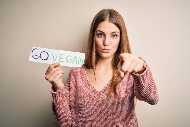 Νεαρή όμορφη κοκκινομάλλα γυναίκα που ζητά veggie τροφίμων κρατώντας χαρτί με vegan μήνυμα δείχνοντας με το δάχτυλο στην κάμερα και σε σας, το χέρι σημάδι, θετική και σίγουρη χειρονομία από το μέτωπο - Φωτογραφία, εικόνα