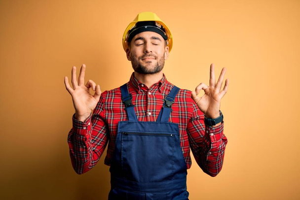 Jeune homme de chantier portant uniforme de construction et casque de sécurité sur fond jaune isolé se détendre et sourire les yeux fermés en faisant geste de méditation avec les doigts. Concept de yoga
. - Photo, image