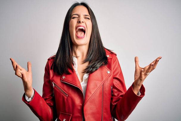 Young brunette woman with blue eyes wearing red casual jacket over white background crazy and mad shouting and yelling with aggressive expression and arms raised. Frustration concept. - Photo, Image