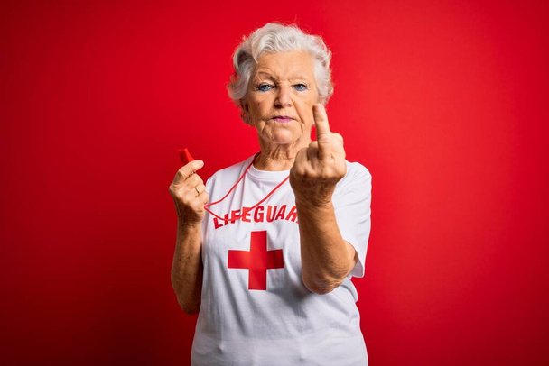 Senior beautiful grey-haired lifeguard woman wearing t-shirt with red cross using whistle Showing middle finger, impolite and rude fuck off expression - Zdjęcie, obraz