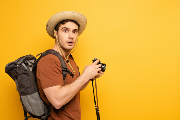 viajante chocado em chapéu com mochila segurando câmera fotográfica no amarelo
 - Foto, Imagem