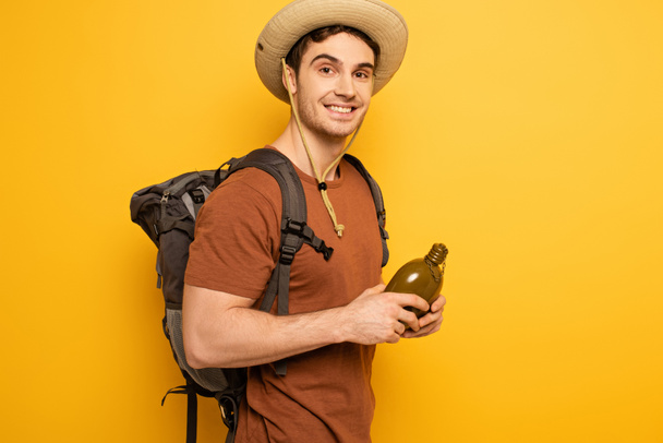 handsome traveler in hat with backpack holding bottle with water on yellow - Photo, Image