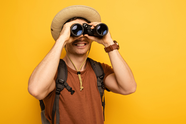 smiling traveler in hat with backpack looking through binoculars  on yellow - Photo, Image