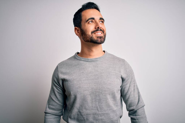 Young handsome man with beard wearing casual sweater standing over white background looking away to side with smile on face, natural expression. Laughing confident. - Photo, Image