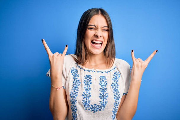 Genç, güzel, esmer, sıradan tişörtler giyen, mavi arka planda dikilmiş çılgın bir ifadeyle elleri havada rock sembolü yapan bir kadın. Müzik yıldızı. Ağır konsept. - Fotoğraf, Görsel
