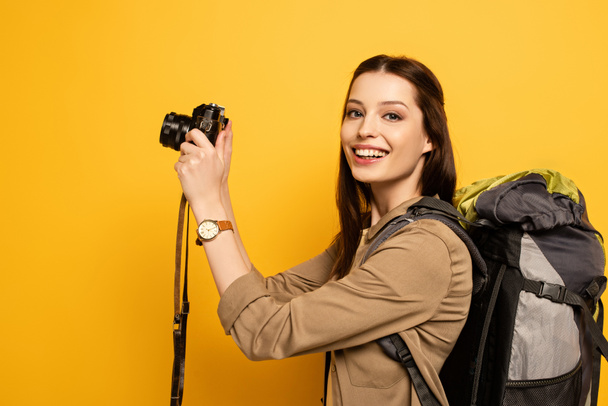 όμορφη χαμογελαστή τουρίστας με σακίδιο κρατώντας φωτογραφική μηχανή φωτογραφία στο κίτρινο  - Φωτογραφία, εικόνα