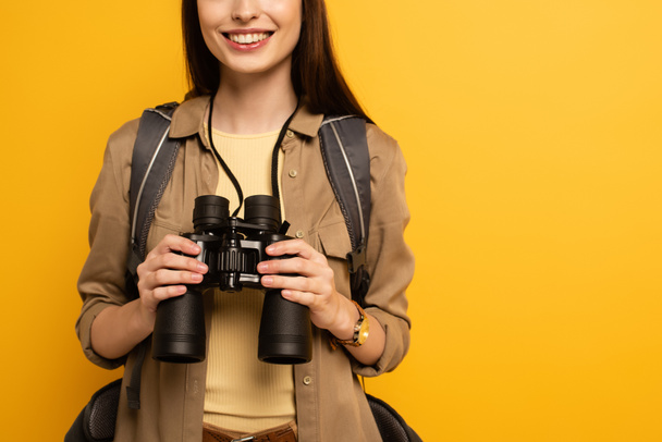 cropped view of traveler with backpack holding binoculars, isolated on yellow  - Фото, зображення