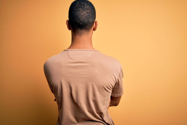 Joven hombre afroamericano guapo usando camiseta casual de pie sobre fondo amarillo de pie hacia atrás mirando hacia otro lado con los brazos cruzados
 - Foto, imagen