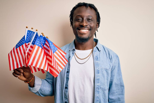 Mladý africký americký vlastenecký muž drží Spojené státy vlajky nad bílým pozadím s veselou tváří stojí a usmívá se s sebevědomým úsměvem ukazuje zuby - Fotografie, Obrázek