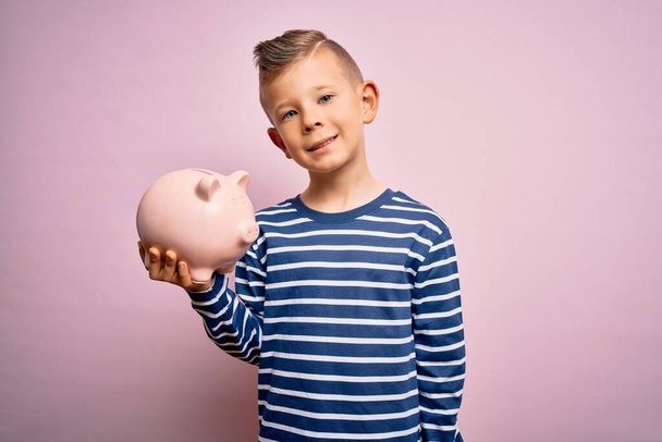 Νεαρό μικρό καυκάσιο παιδί τραπεζικά χρήματα σε κουμπαρά ως εξοικονόμηση πλούτου πάνω από ροζ φόντο με ένα χαρούμενο πρόσωπο στέκεται και χαμογελά με αυτοπεποίθηση χαμόγελο δείχνει τα δόντια - Φωτογραφία, εικόνα
