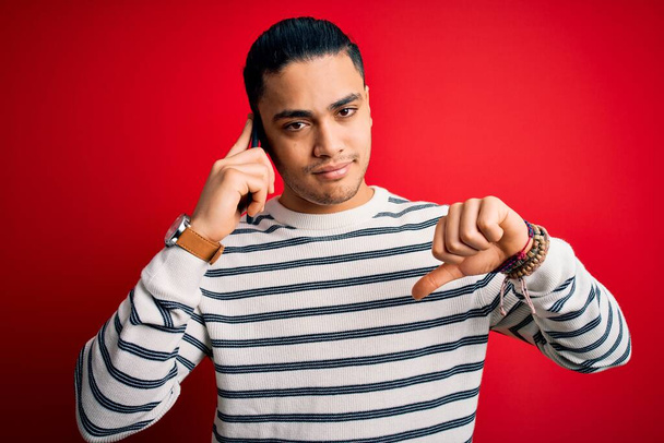 Brazilian man having conversation talking on the smartphone over isolated red background with angry face, negative sign showing dislike with thumbs down, rejection concept - Foto, afbeelding