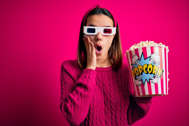Νεαρό όμορφο κορίτσι βλέποντας ταινία χρησιμοποιώντας 3d γυαλιά τρώει κουτί με ποπ κορν φοβάται και σοκαρισμένος, έκπληξη και κατάπληκτος έκφραση με τα χέρια στο πρόσωπο - Φωτογραφία, εικόνα