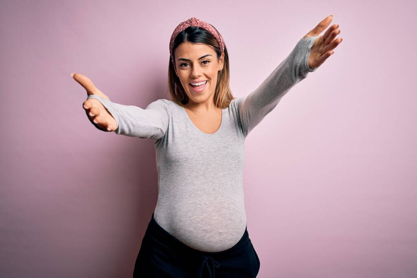 Νεαρή όμορφη μελαχρινή γυναίκα έγκυος περιμένει μωρό πάνω από απομονωμένο ροζ φόντο κοιτάζοντας την κάμερα χαμογελώντας με ανοιχτές αγκάλες για αγκαλιά. Χαρούμενη έκφραση που αγκαλιάζει την ευτυχία. - Φωτογραφία, εικόνα