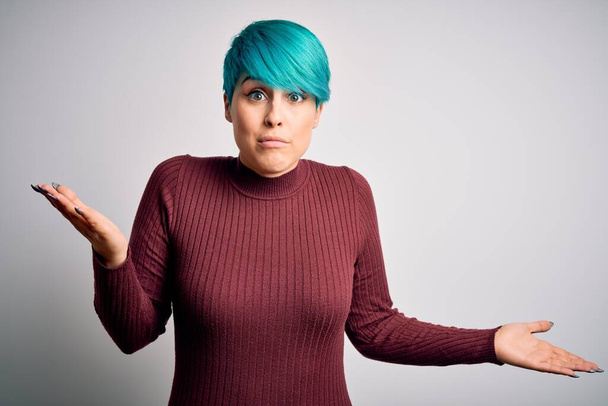 Young beautiful woman with blue fashion hair wearing casual sweater over white background clueless and confused expression with arms and hands raised. Doubt concept. - Fotografie, Obrázek