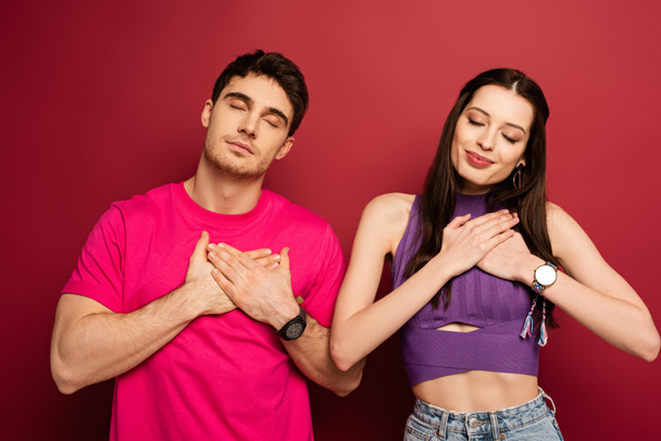 positive young couple with closed eyes holding hands on hearts on red - Photo, image