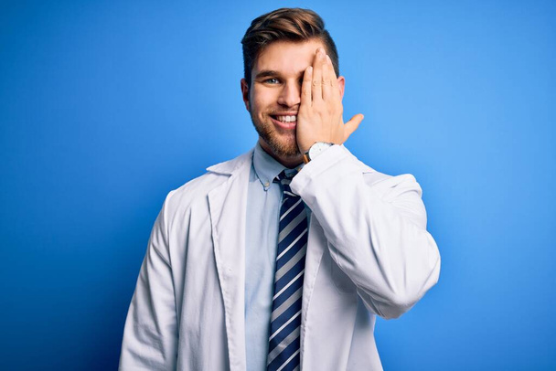 Young blond therapist man with beard and blue eyes wearing coat and tie over background covering one eye with hand, confident smile on face and surprise emotion. - Foto, imagen