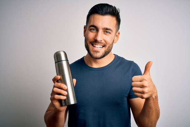 Young handsome tourist man drinking thermo with water over isolated white background happy with big smile doing ok sign, thumb up with fingers, excellent sign - Foto, imagen