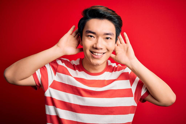 Nuori komea kiinalainen mies päällään rento raidallinen t-paita seisoo yli punainen tausta yrittää kuulla molemmat kädet korva ele, utelias juoruja. Kuulo-ongelma, kuuro
 - Valokuva, kuva