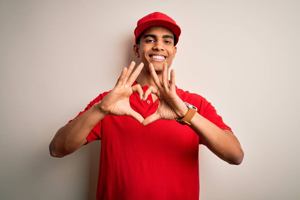 カジュアルなポロを身に着けている若いハンサムなアフリカ系アメリカ人男性と手で心のシンボルと形状を示す愛に笑みを浮かべて赤い背景にキャップ。ロマン主義. - 写真・画像