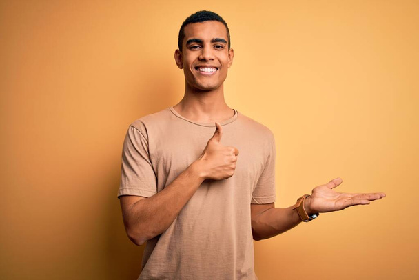 Young handsome african american man wearing casual t-shirt standing over yellow background Showing palm hand and doing ok gesture with thumbs up, smiling happy and cheerful - Fotografie, Obrázek