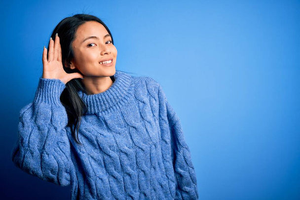 Fiatal, gyönyörű, kínai nő alkalmi pulóvert visel elszigetelt kék háttér felett mosolyogva, fültől fülig hallgatva egy pletykát vagy pletykát. Süketség fogalma. - Fotó, kép