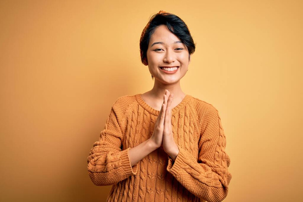 Nuori kaunis aasialainen tyttö päällään rento pusero ja diadem seisoo keltainen tausta rukoilee kädet yhdessä pyytäen anteeksiantoa hymyillen luottavainen
. - Valokuva, kuva