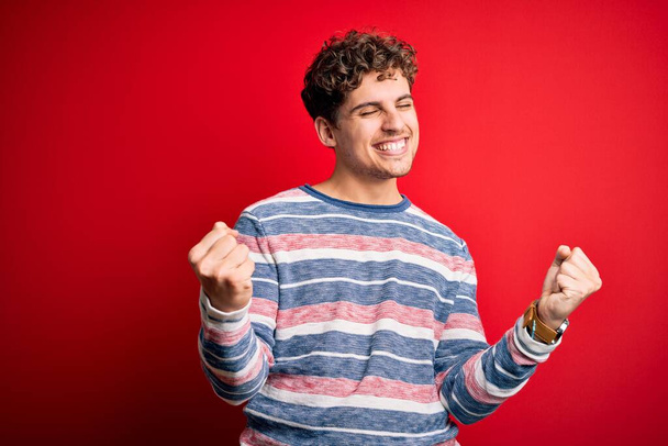 Young blond handsome man with curly hair wearing striped sweater over red background very happy and excited doing winner gesture with arms raised, smiling and screaming for success. Celebration concept. - Photo, Image