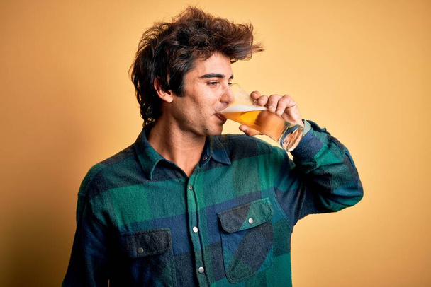 幸せな笑顔の若いハンサムな男。孤立した黄色の背景の上にビールの顔を飲むガラスに笑顔で立って - 写真・画像