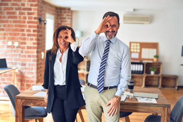 Twee werknemers van middelbare leeftijd die samen werken in een vergadering op kantoor doen ok gebaar met de hand glimlachen, oog kijken door vingers met gelukkig gezicht. - Foto, afbeelding