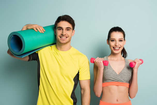 happy athletic couple holding fitness mat and dumbbells on blue - Photo, Image