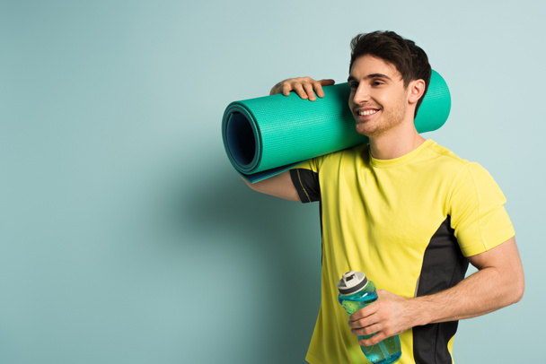 smiling muscular sportsman in yellow t-shirt holding fitness mat and sports bottle with water on blue - Foto, Imagen