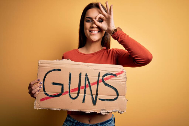 Młoda piękna aktywistka prosi o pokój trzymając sztandar z stop broni wiadomość ze szczęśliwą twarzą uśmiech robi ok znak z ręki na oko patrząc przez palce - Zdjęcie, obraz
