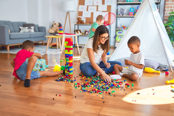 Joven hermosa maestra y niños pequeños jugando con bloques de construcción alrededor de un montón de juguetes en el jardín de infantes
 - Foto, imagen