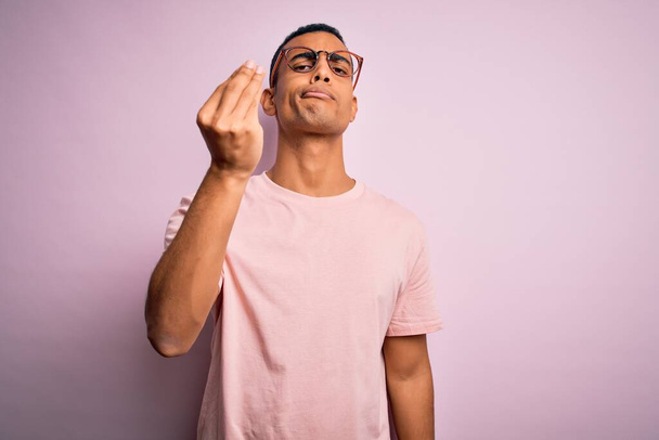 Schöner afrikanisch-amerikanischer Mann mit lässigem T-Shirt und Brille vor rosa Hintergrund Italienische Geste mit selbstbewusstem Ausdruck an Händen und Fingern - Foto, Bild