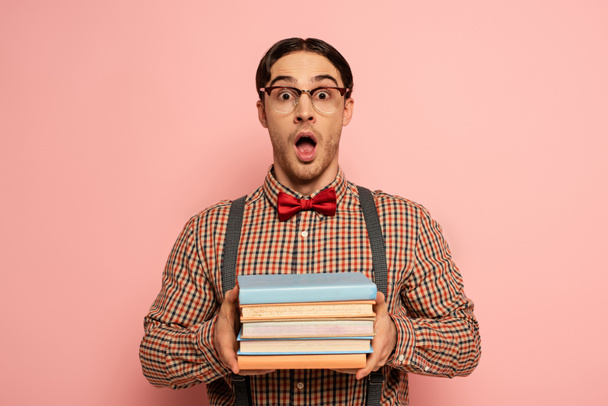 shocked male nerd with open mouth in eyeglasses holding books on pink - Fotografie, Obrázek