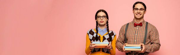 panoramic shot of couple of happy nerds in eyeglasses holding books, Isolated on pink - Foto, afbeelding