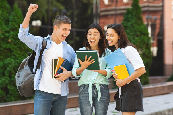 Радостные студенты университета читают результаты экзаменов, стоя в кампусе, празднуя успех
 - Фото, изображение
