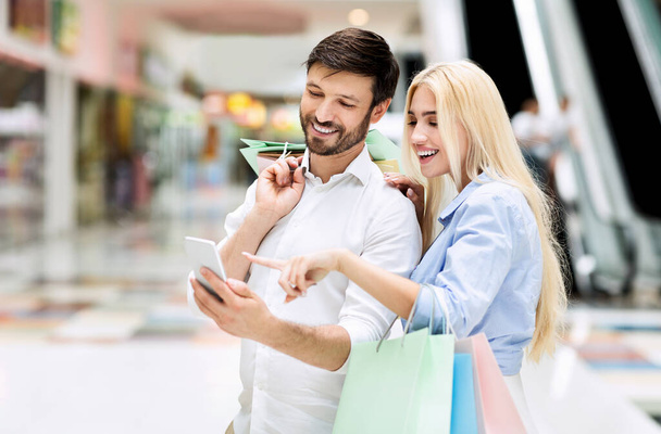 Σύζυγοι που χρησιμοποιούν Smartphone Ψάχνοντας τις πωλήσεις και τις προσφορές στέκεται στο εμπορικό κέντρο - Φωτογραφία, εικόνα
