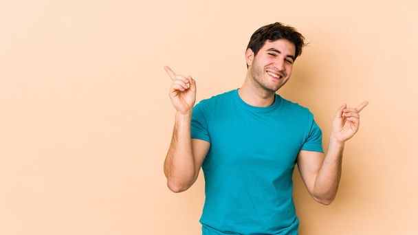 Young man isolated on beige background pointing to different copy spaces, choosing one of them, showing with finger. - Foto, Imagen