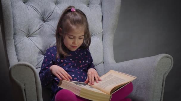 urocza dziewczynka patrzy przez książkę na krześle - Materiał filmowy, wideo