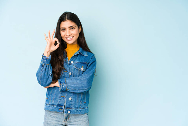 Young indian woman isolated on blue background winks an eye and holds an okay gesture with hand. - Photo, Image