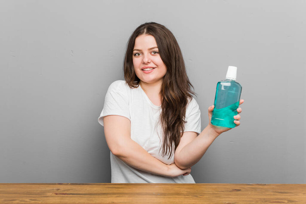 Young plus size curvy woman holding a mouthwash smiling confident with crossed arms. - Photo, Image