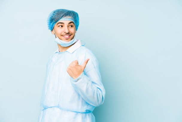 Junger arabischer Chirurg isoliert gegen auf blauem Hintergrund zeigt mit dem Daumen Finger weg, lachend und unbeschwert. - Foto, Bild