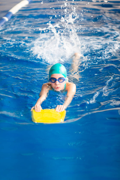 Cute little girl swimmer training in a swimming pool in glasses with a swimming kickboard. Swimming sport for kids concept - Фото, зображення