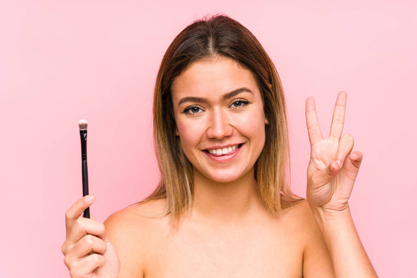 Young caucasian woman holding a eyebrush isolated showing victory sign and smiling broadly. - Foto, Imagem