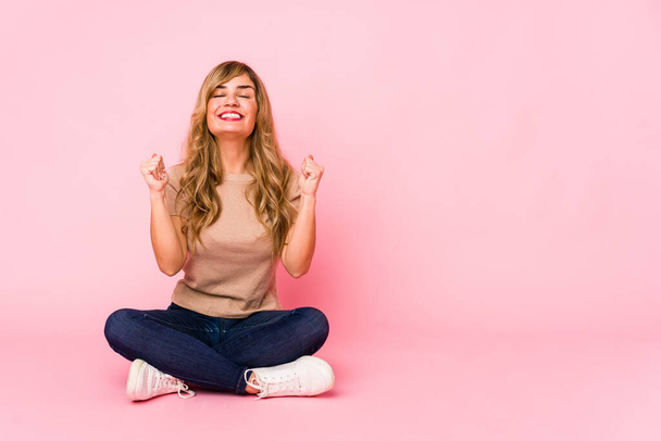 Молодая блондинка-кавказка сидит на розовой студии, поднимая кулаки, чувствуя себя счастливой и успешной. Идея победы
. - Фото, изображение