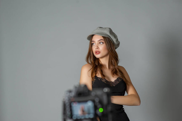 Czarująca młoda kobieta nosi modną sukienkę pozującą z trzepoczącymi włosami na szarej ścianie.Dziewczyna wysunięta do przodu ręka w czarnej sukience i szarej czapce.Dziewczyna jest fotografowana na aparacie lub ja kręcę film - Zdjęcie, obraz